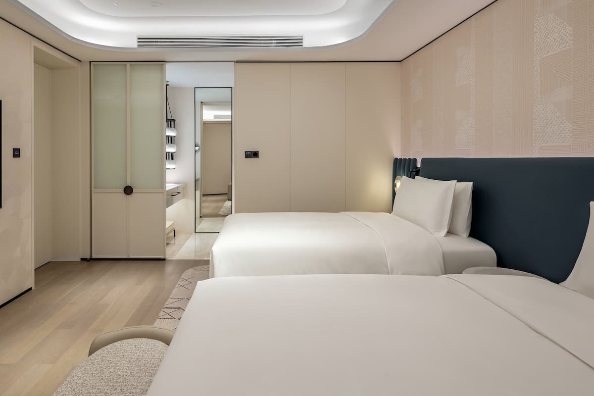 A modern hotel room with a flat-screen TV, two beds, a walk-in closet, and a ceiling light fixture.