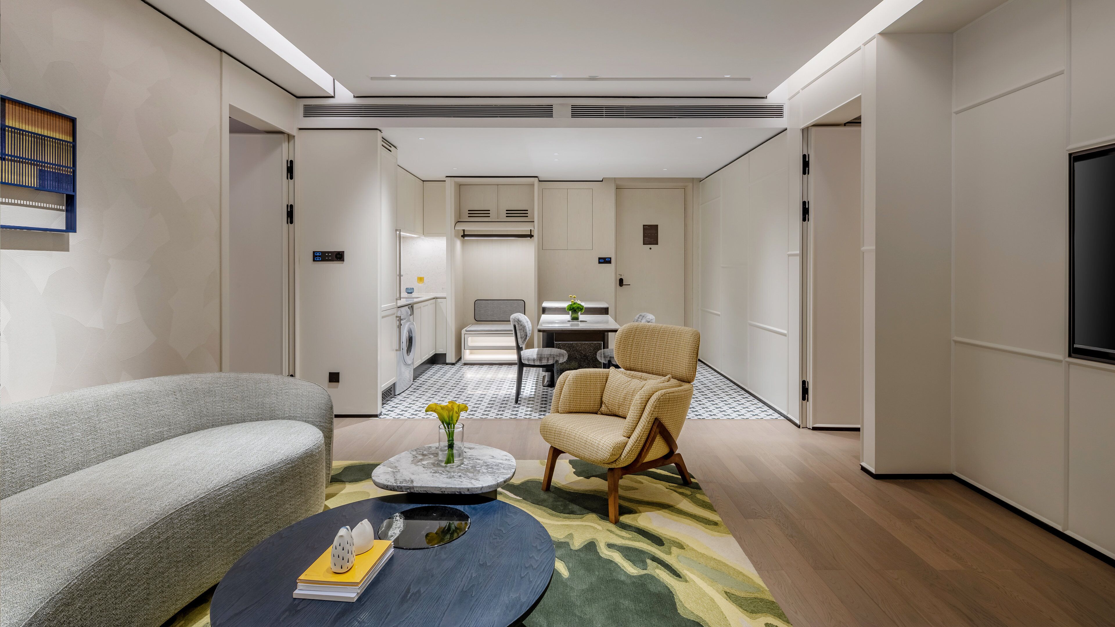 A modern hotel room with a sofa, armchair, dining table, and a flat-screen TV.