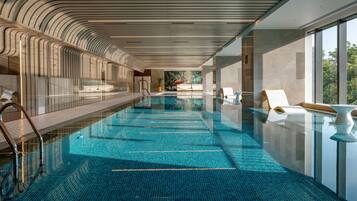 A modern indoor swimming pool with a tiled floor, lounge chairs, and a view of trees through large windows.