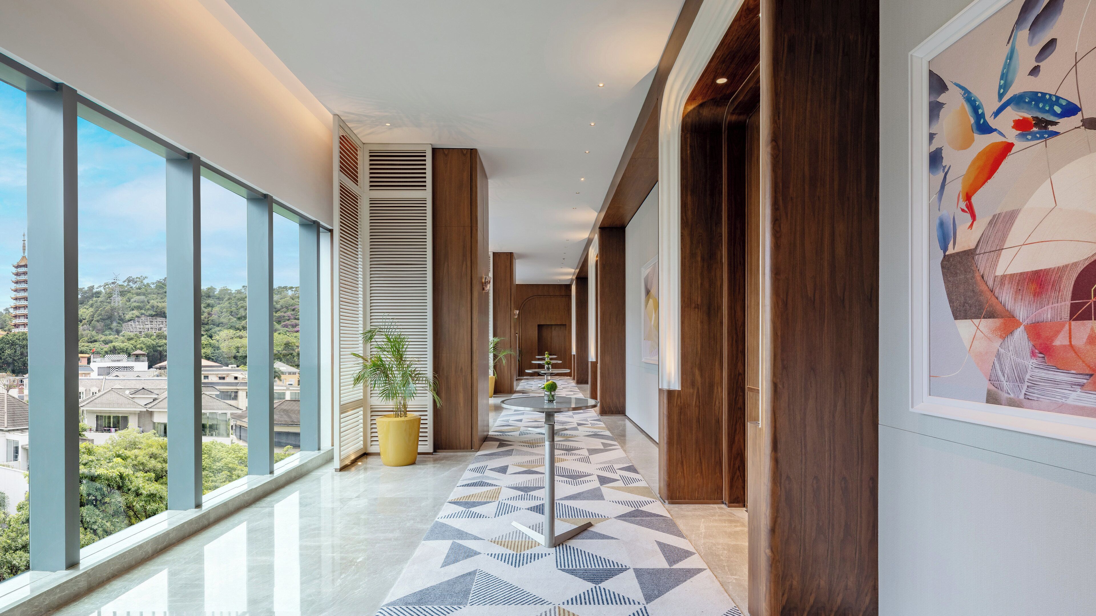 A modern hotel corridor with large windows, a geometric patterned floor, and a view of buildings and greenery.