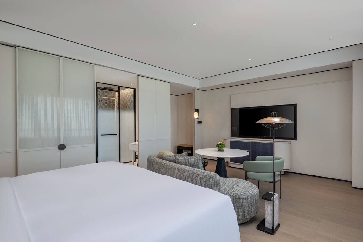 A modern hotel room with a large bed, a sofa, a small round table, and a TV mounted on the wall.