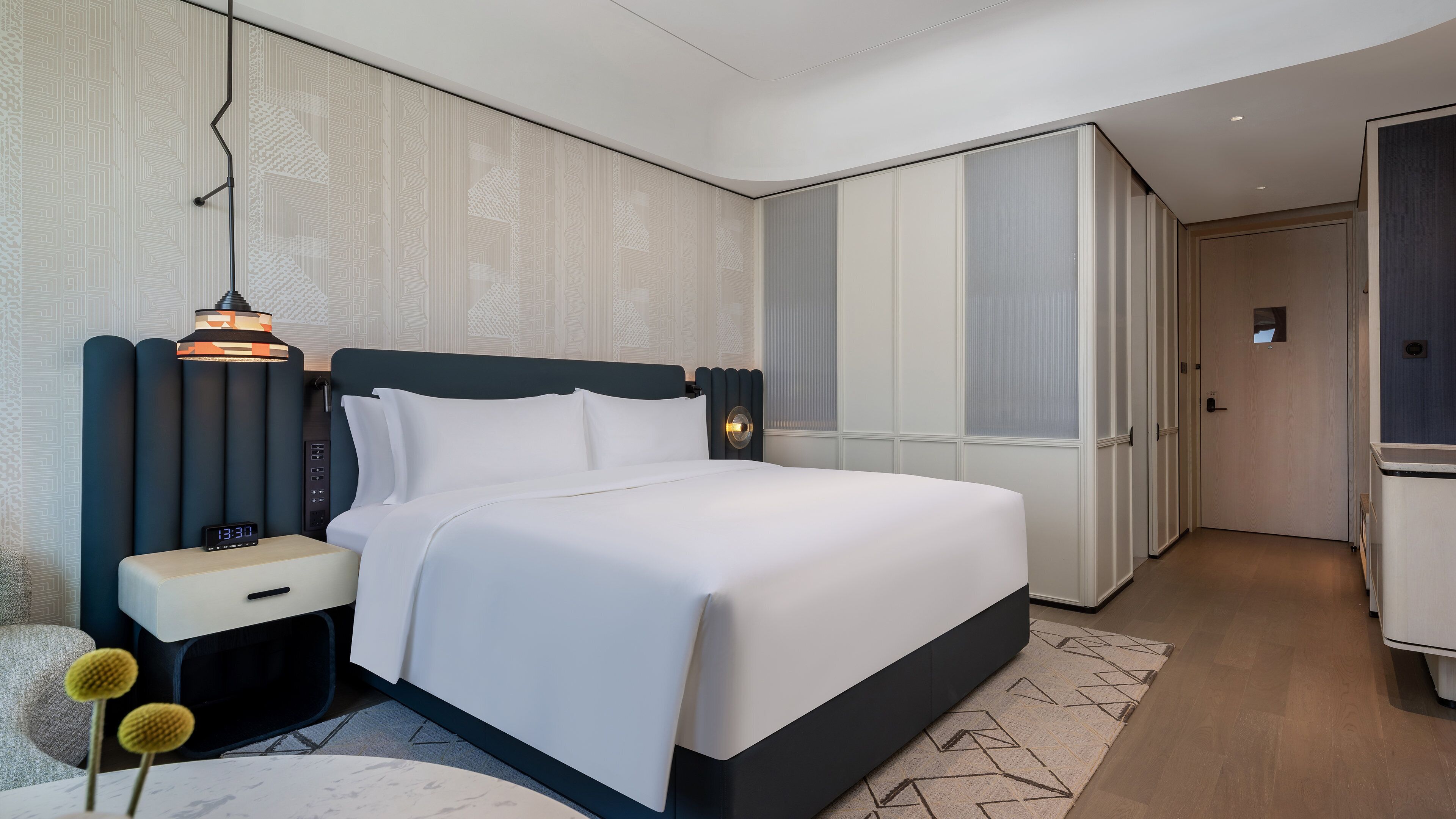 Ein modernes Hotelzimmer mit Bett, Nachttisch, Lampe, Schrank und einem kleinen Tisch mit Dekoration.