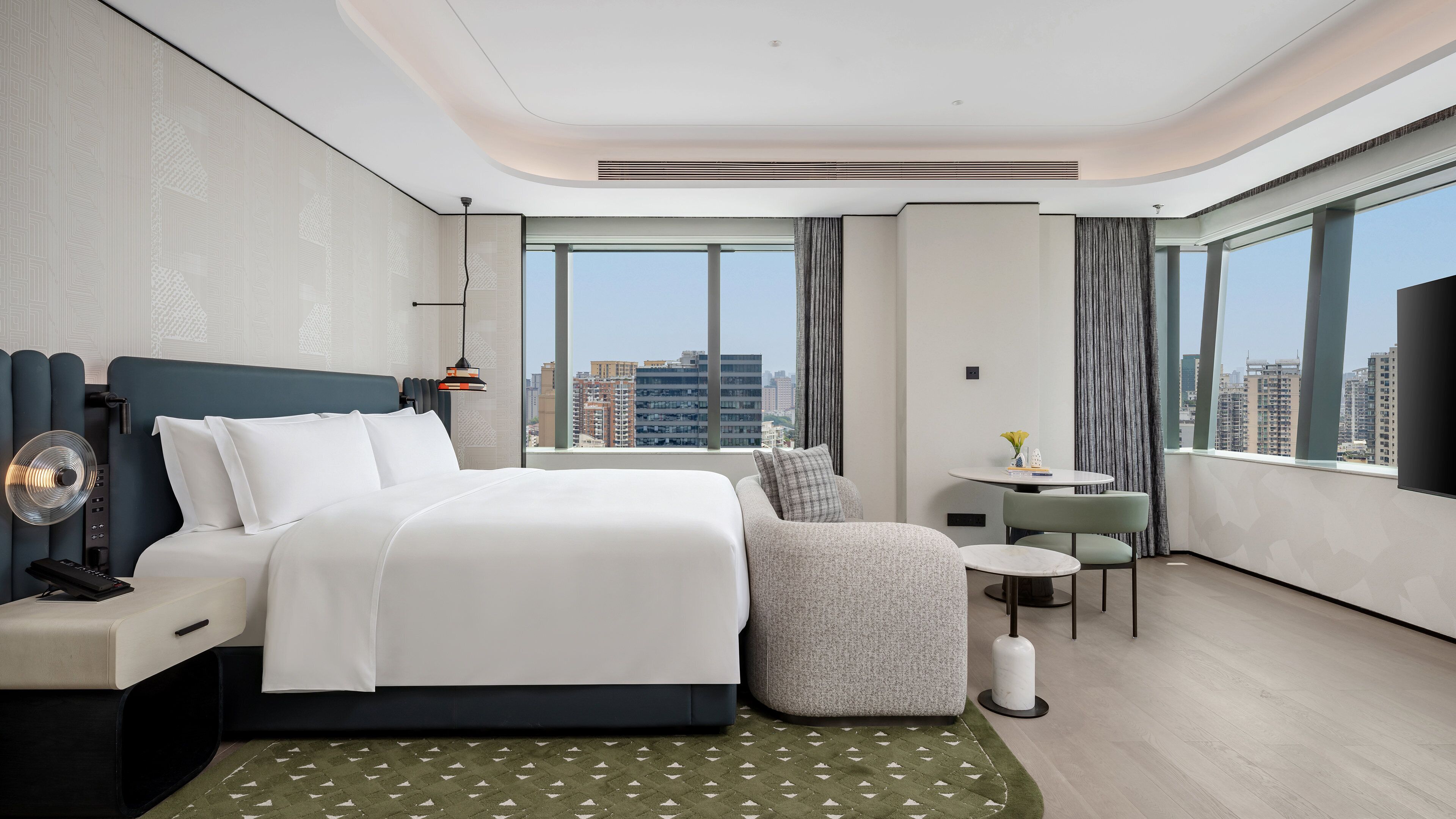 A modern hotel room with a large bed, a desk, a chair, a small table, and a view of the city skyline.