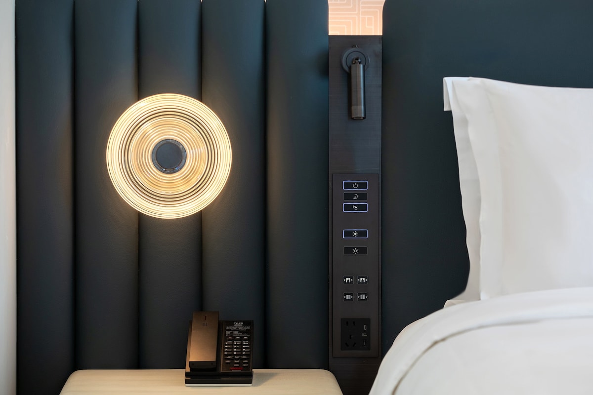 A modern hotel room with a circular light fixture, a control panel with multiple buttons, and a bedside table with a remote control.