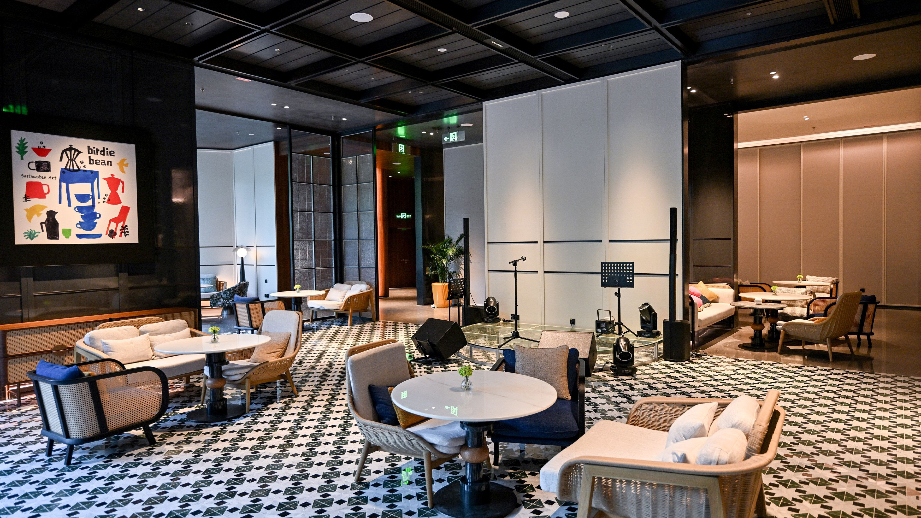 Un moderno vestíbulo de hotel con zonas de estar, un escenario y una pantalla de proyección que muestra arte.