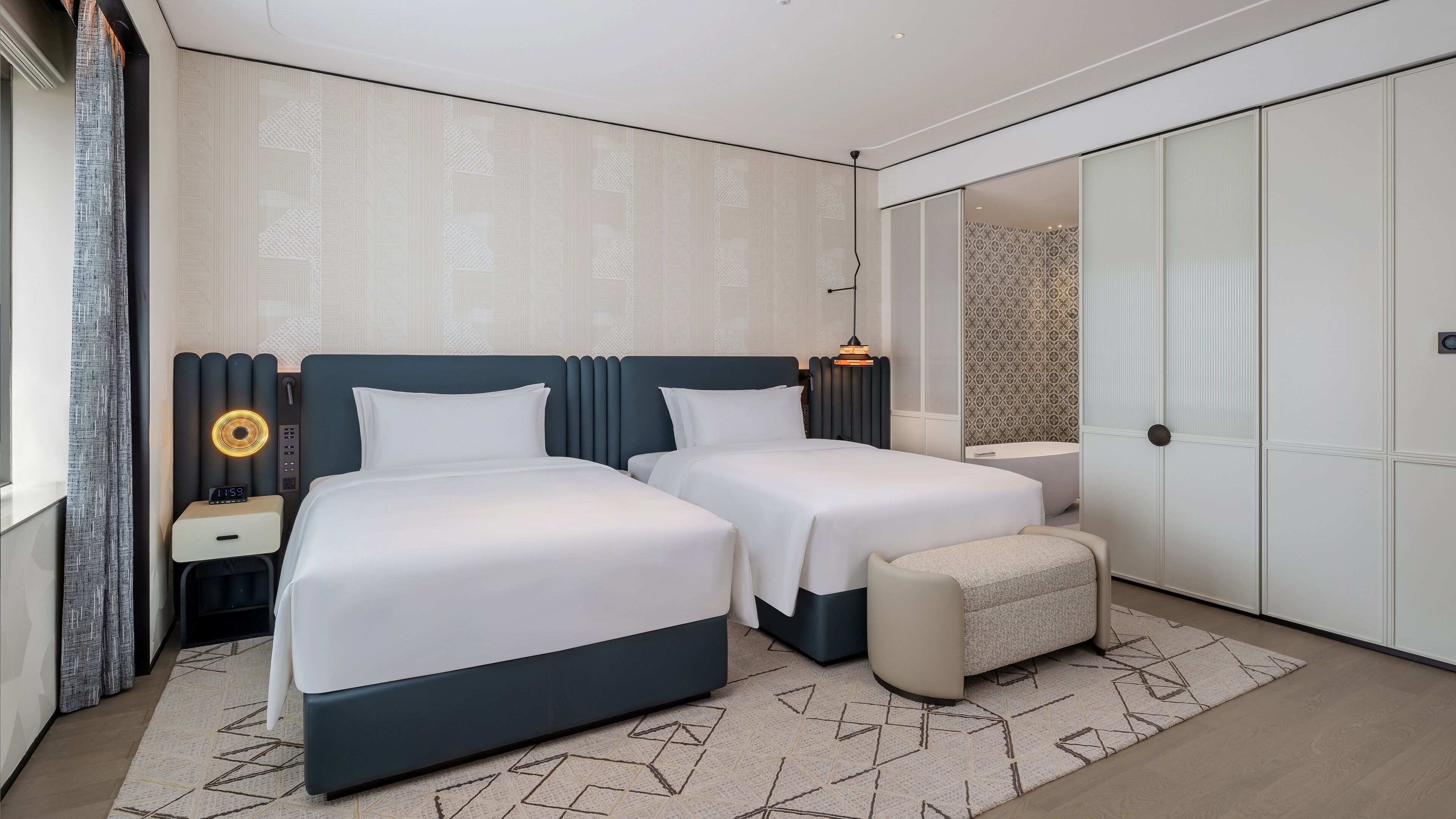 Una habitación de hotel moderna con dos camas, una mesita de noche con lámpara, un taburete pequeño y un armario grande.