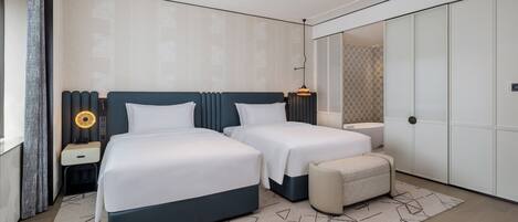 Una habitación de hotel moderna con dos camas, una mesita de noche con lámpara, un taburete pequeño y un armario grande.