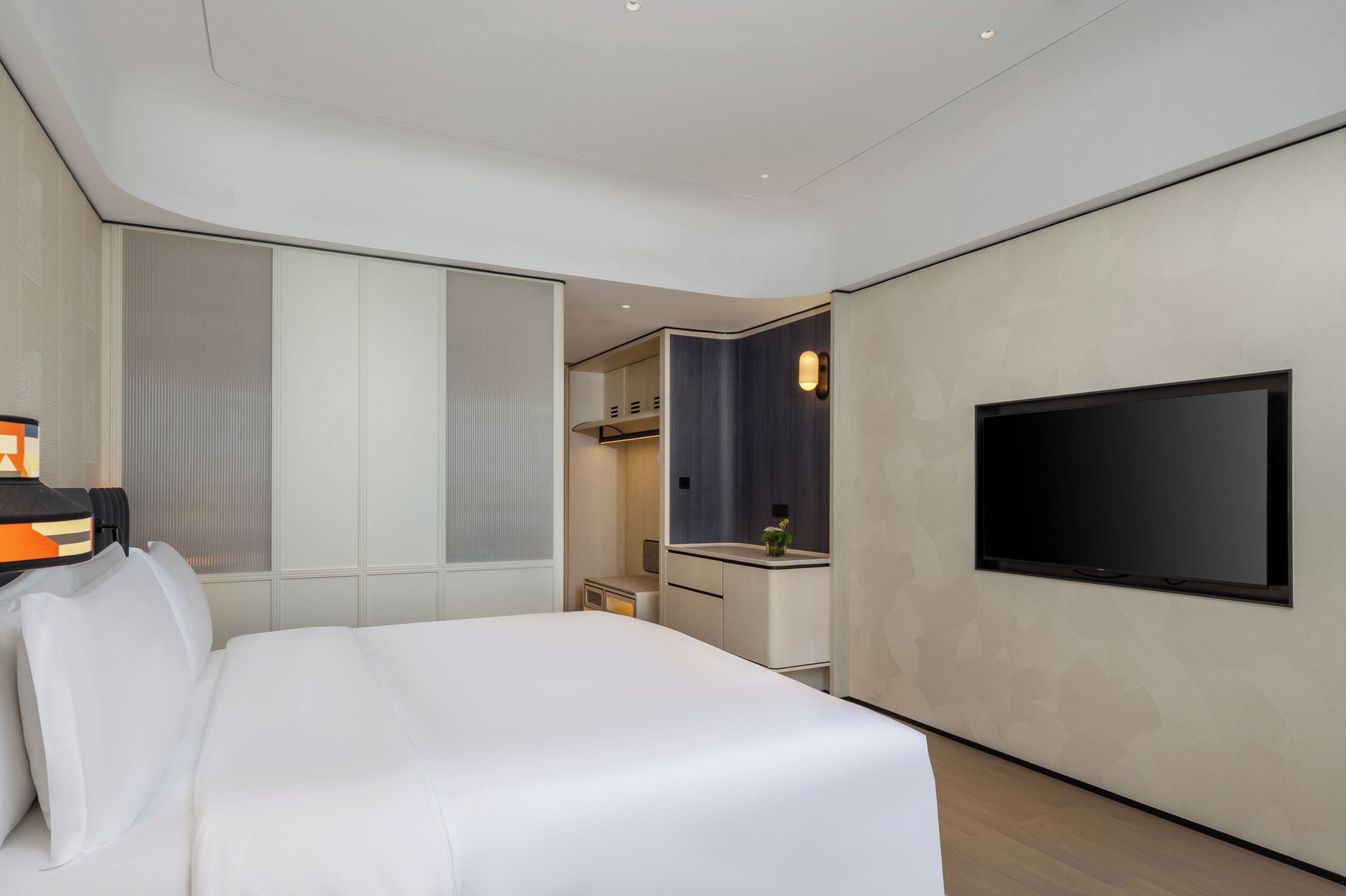 A modern hotel room with a large bed, a flat-screen TV mounted on the wall, and a small kitchenette area.