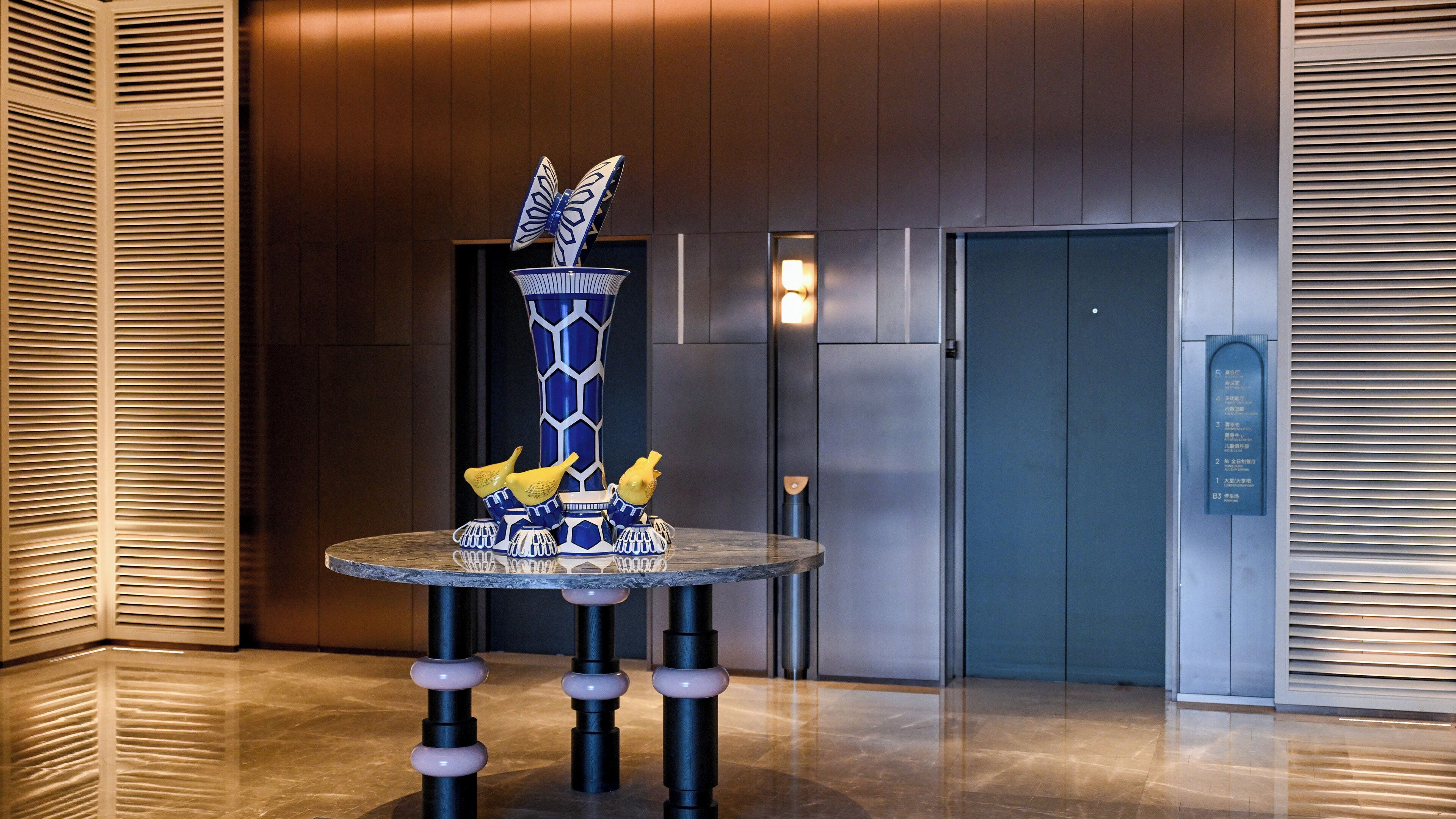 Un hall d’entrée moderne agrémenté d’un vase bleu et blanc original, d’une porte donnant accès à un ascenseur et d’un mur orné de lignes horizontales.
