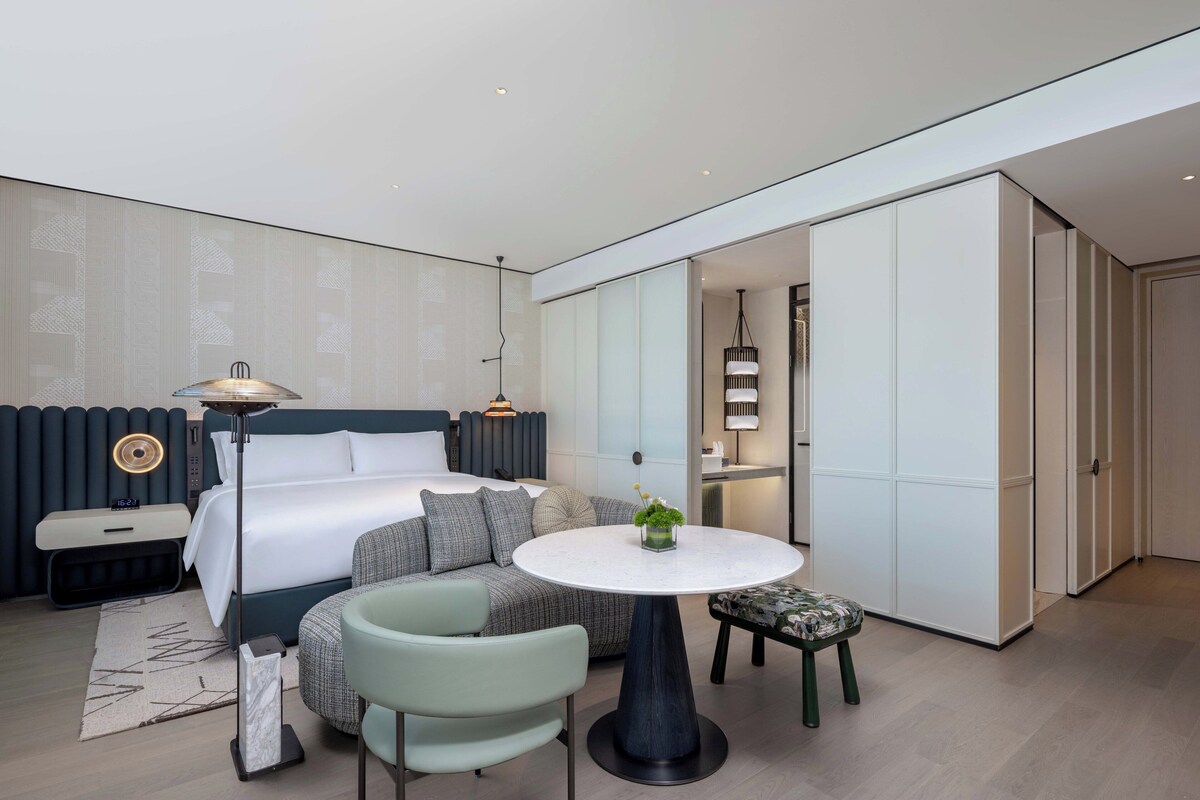 A modern hotel room with a bed, a sofa, a round table, and a small stool.