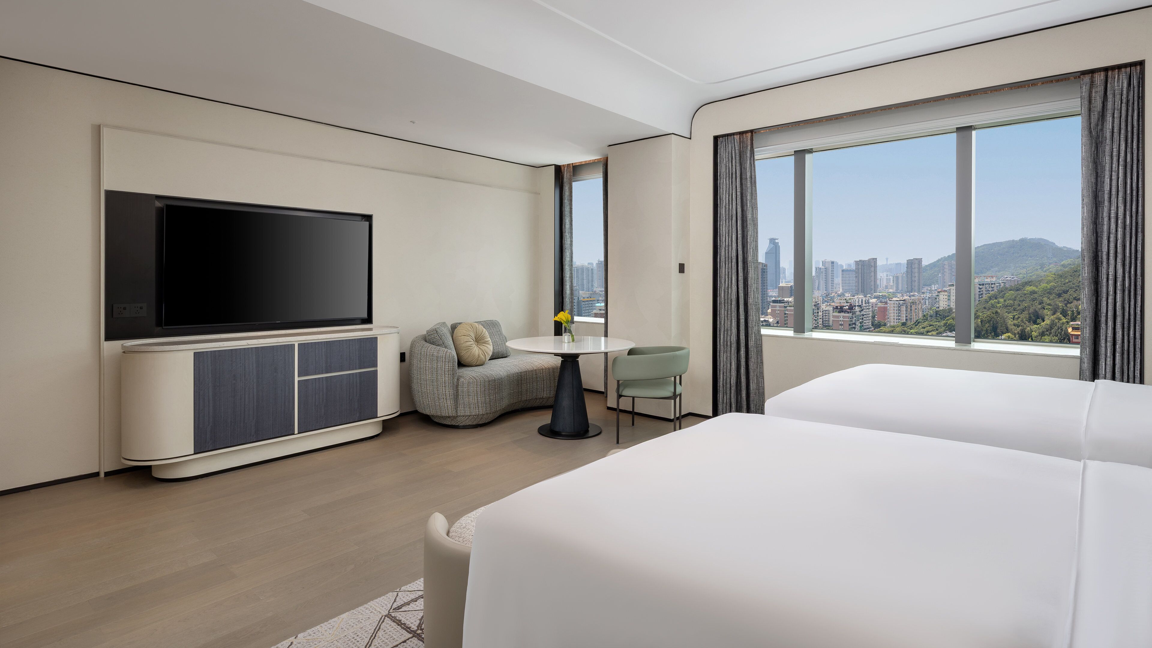 Habitación de hotel moderna con televisor de pantalla plana, una pequeña mesa de comedor, un sofá y un ventanal con vistas a la ciudad.