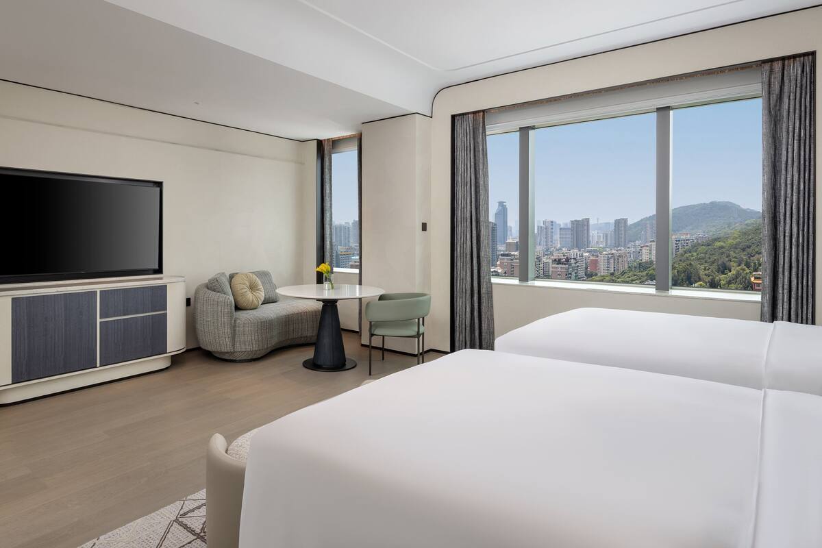 A modern hotel room with a flat-screen TV, a small dining table, a sofa, and a large window offering a city view.