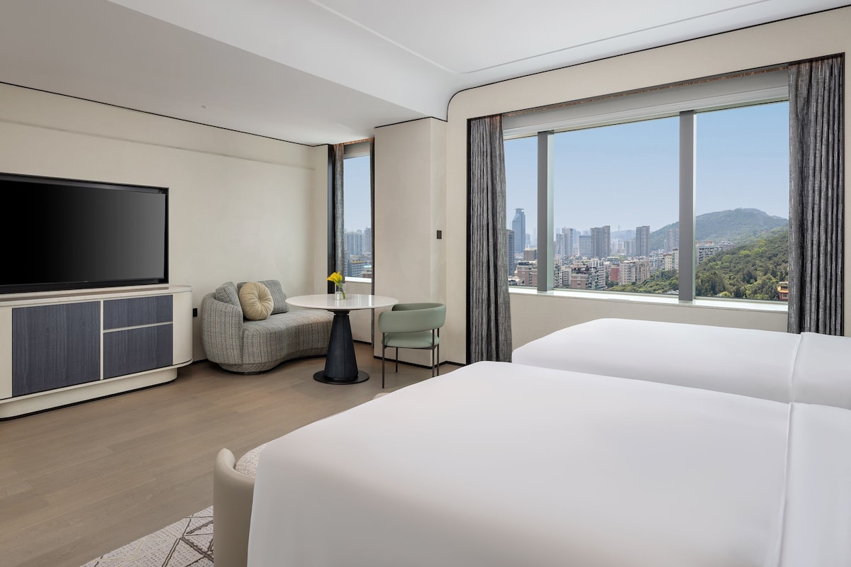 A modern hotel room with a flat-screen TV, a small dining table, a sofa, and a large window offering a city view.