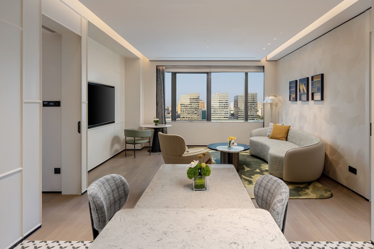 A modern living room with a dining table, sofa, and chairs, large windows with a city view, and a flat-screen TV.