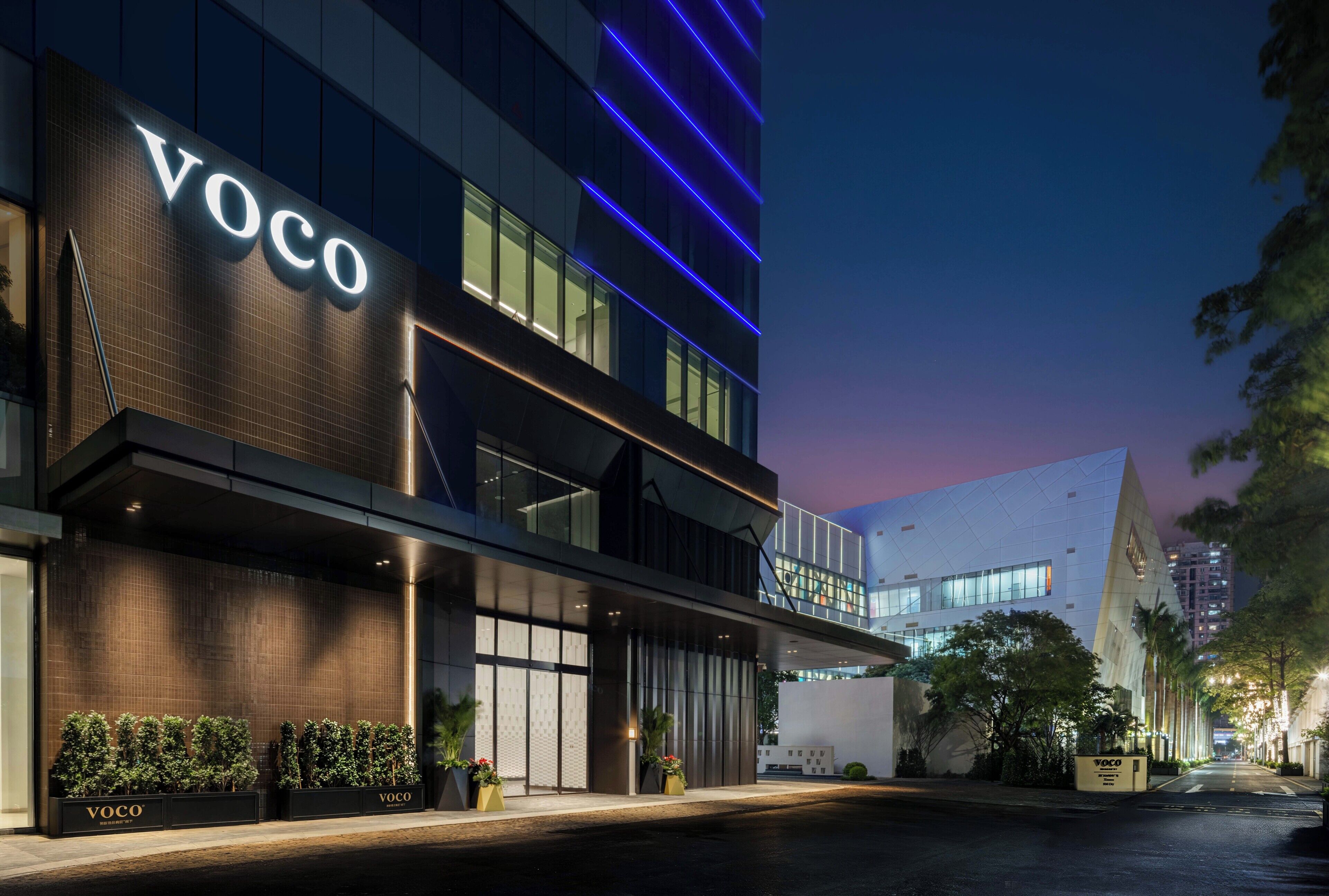 A modern building with a prominent 'VOCO' sign, a covered entrance, and a well-lit facade.