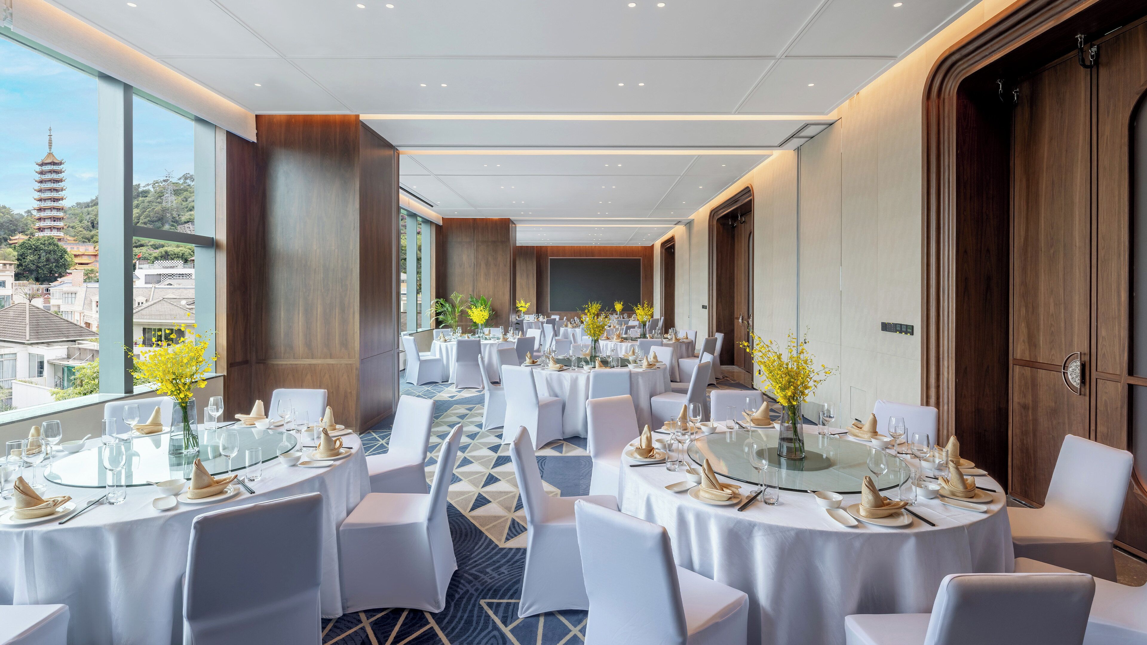 Un salón de banquetes con mesas redondas, sillas blancas y vistas a una pagoda a través de amplios ventanales.