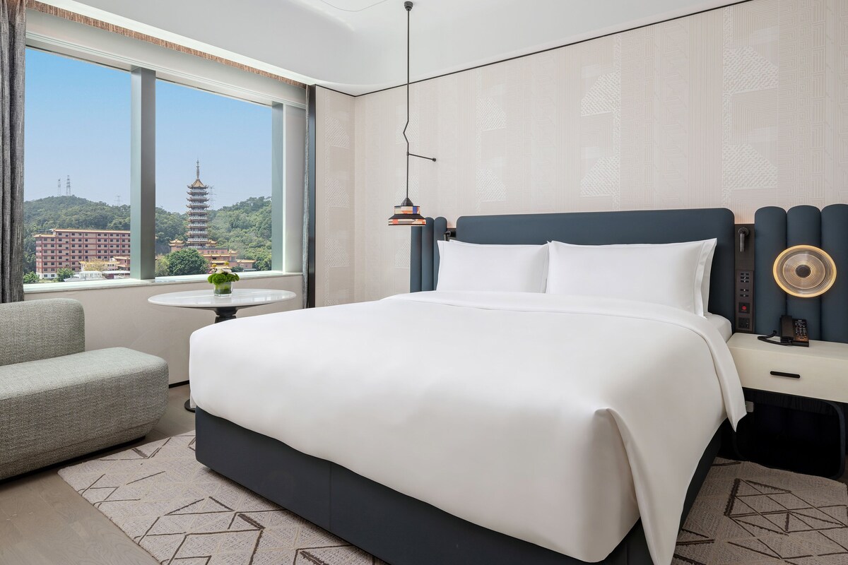 Habitación de hotel con cama, mesita de noche, lámpara, silla y vista a una pagoda a través de una ventana.