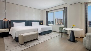Habitación de hotel con dos camas, una mesita con un florero, una silla, una mesita de noche con un teléfono y vistas a edificios.