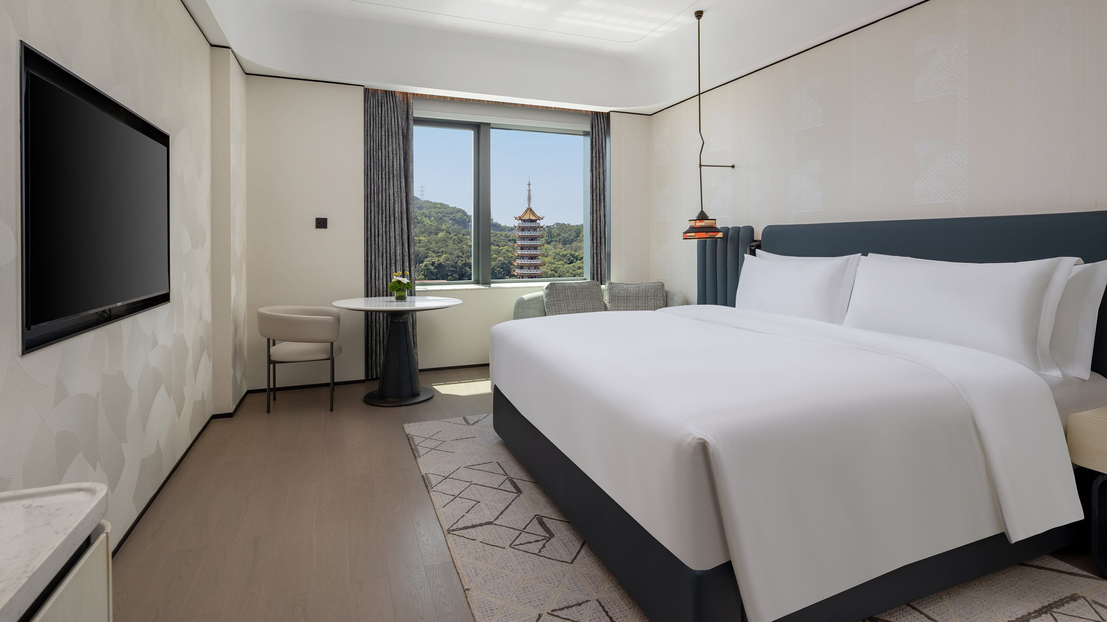 A hotel room with a bed, a TV, a small table with a chair, and a window with a view of a pagoda.