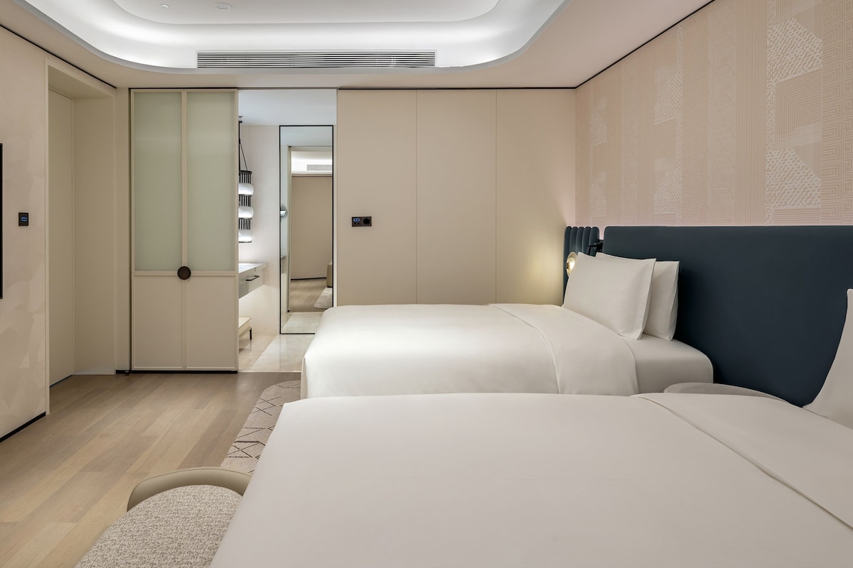 A modern hotel room with a flat-screen TV, two beds, a walk-in closet, and a ceiling light fixture.