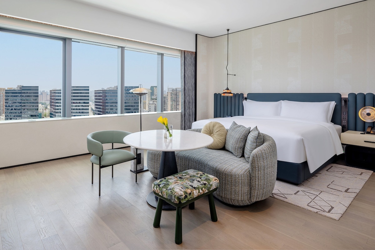 A modern hotel room with a large window, a round table, a sofa, and a bed.