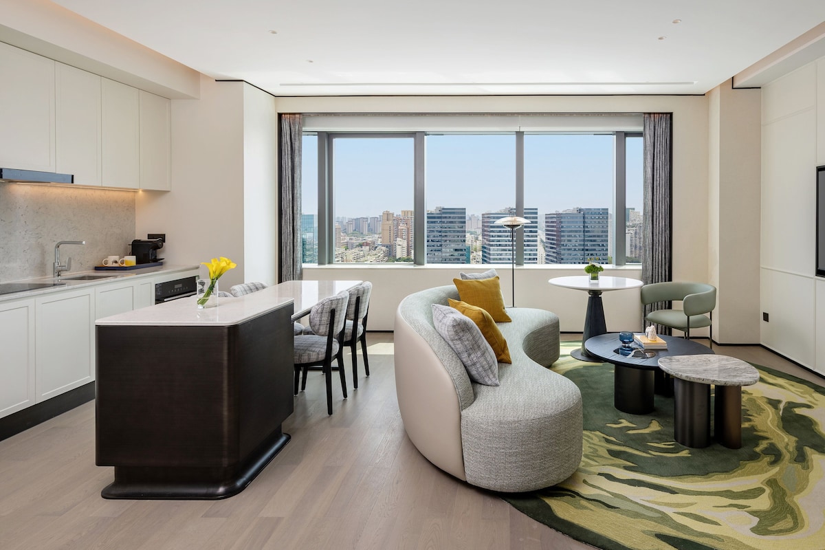 A modern living room with a kitchen area, a large window with a city view, a flat-screen TV, and a round coffee table.