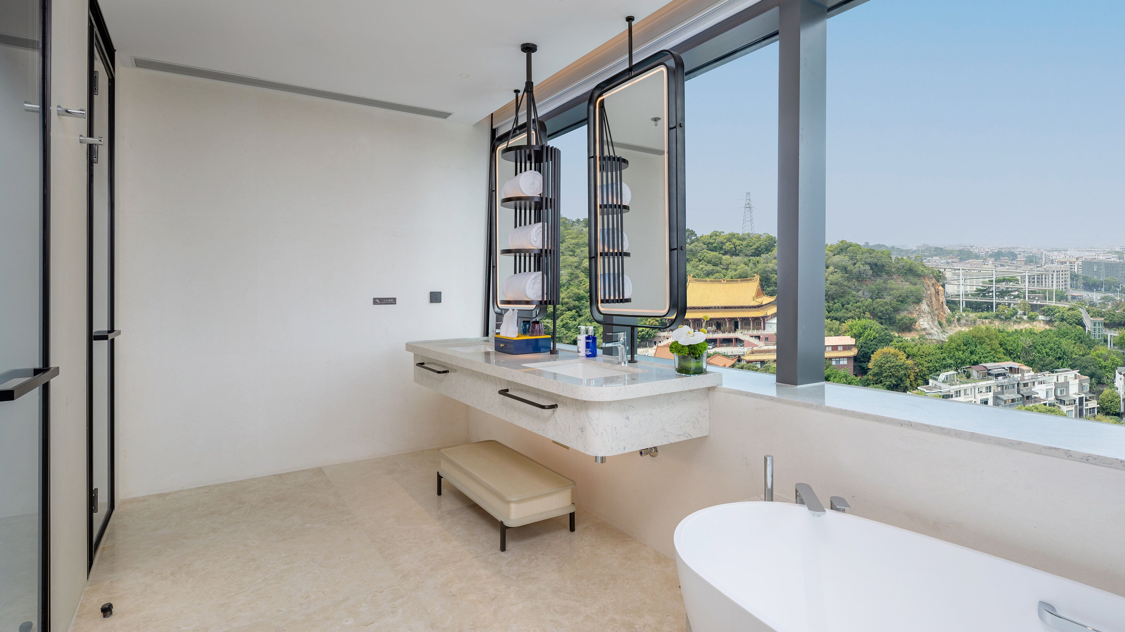 Un baño moderno con una bañera, un tocador con espejo y vistas a un paisaje urbano a través de amplias ventanas.