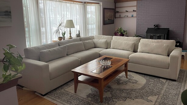 A living room with a beige sectional sofa, a wooden coffee table, and a patterned rug. There is a window with sheer curtains, a lamp, and a small potted plant on the windowsill. - Spacious 3BR/2Bath Home - Retreat w/ Modern Amenities (Harper Woods)