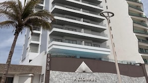 Un hotel moderno e alto con un logo ben visibile alla base, numerosi balconi e una palma in primo piano.