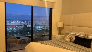 Una camera da letto con un letto grande, un comodino, una lampada, una sedia e vista sulla città attraverso una porta scorrevole.