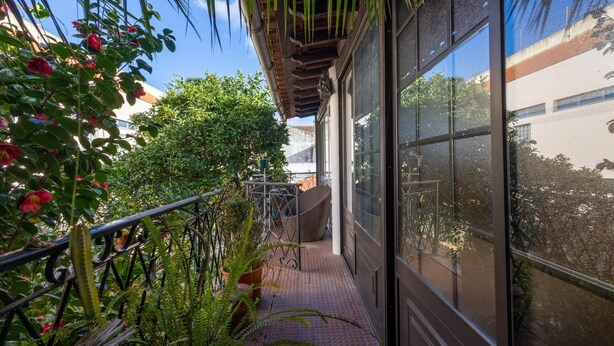 A narrow outdoor corridor with a metal railing, potted plants, and a view of a building through large windows.