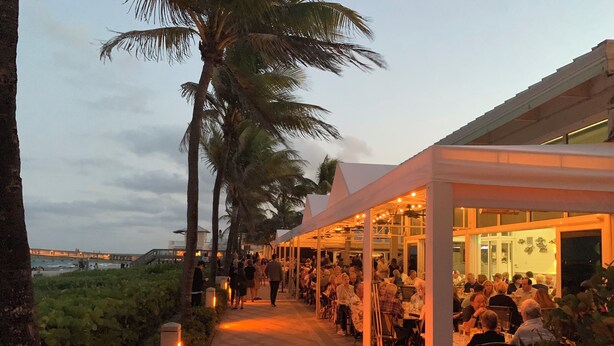 A covered outdoor dining area with tables and chairs, palm trees, and a view of the ocean. - Deerfield Beach Guest Studio (DEERFIELD BEACH)