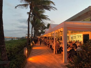 A covered outdoor dining area with tables and chairs, palm trees, and a view of the ocean.