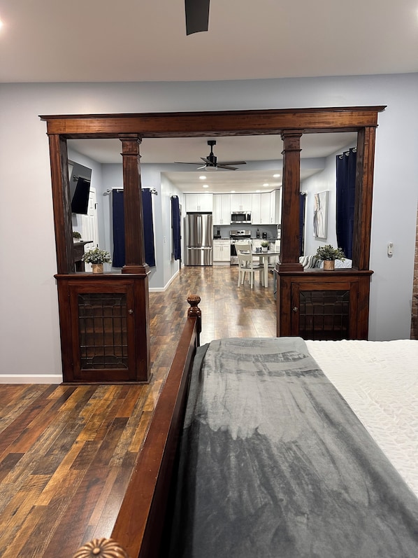 Un dormitorio con una cama con dosel de madera, vista a una cocina con armarios blancos, un refrigerador y un microondas.