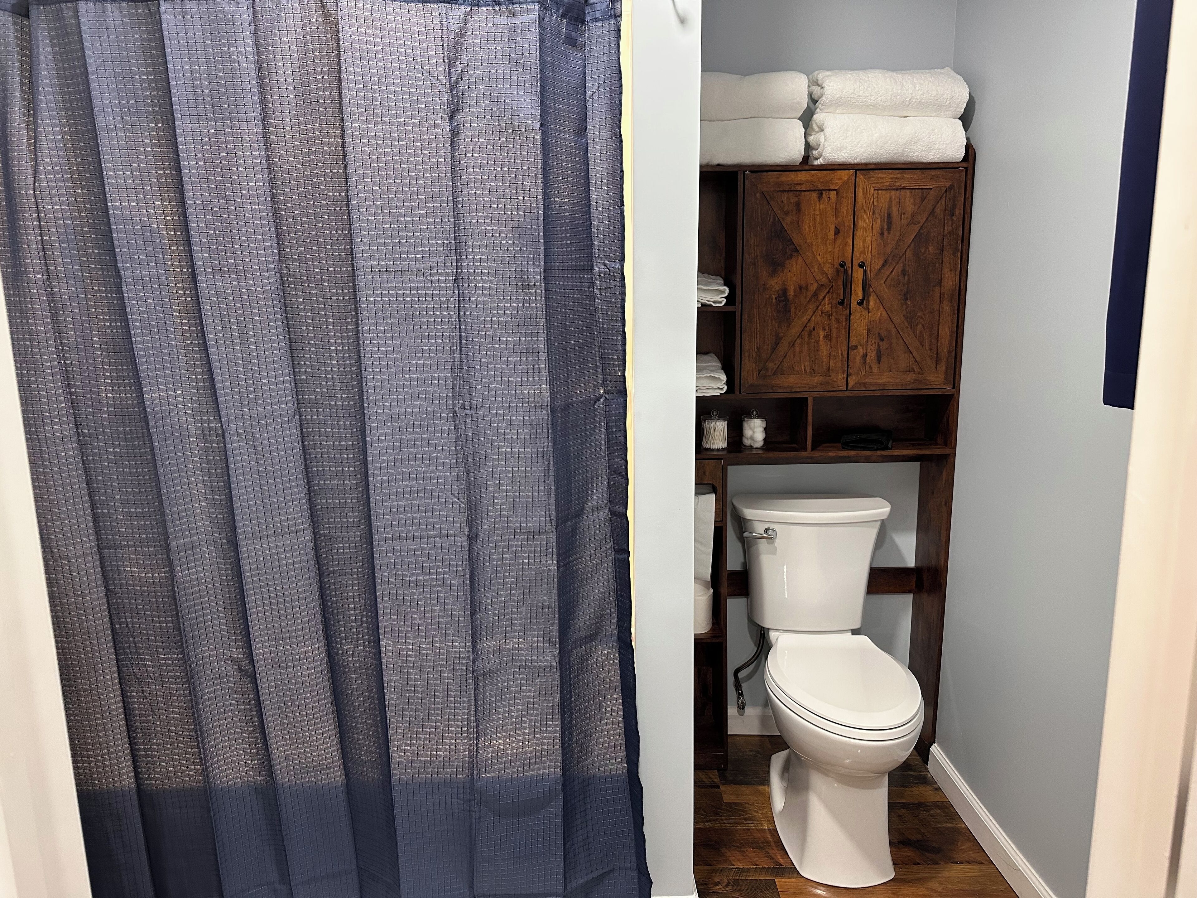 A bathroom with a toilet, a wooden cabinet with towels, and a blue shower curtain.