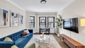 A living room with a blue sofa, a coffee table, a TV, and a dining table. - Beautifully Remodeled 2B/2BA Hyde Park (Chicago)