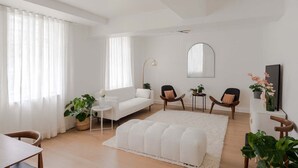 Un salon moderne avec un canapé blanc, deux fauteuils, une table basse et un grand miroir. On y trouve également des plantes en pot et un tapis blanc sur le sol.