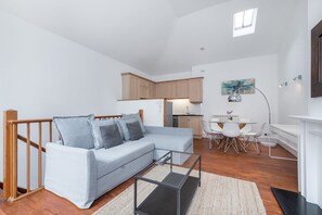 Een moderne woonkamer met een grijze modulaire bank, een glazen salontafel en een eethoek met witte stoelen en een kleine tafel.