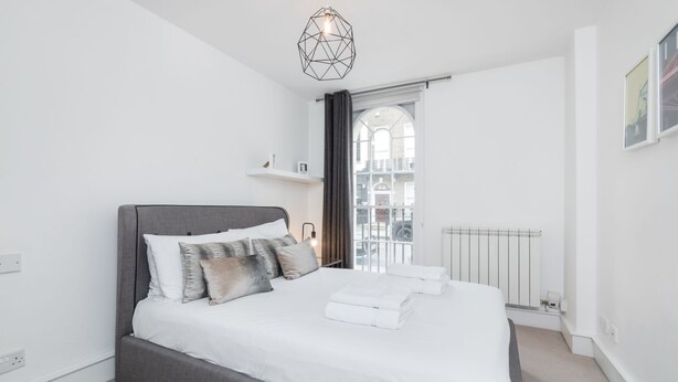 A modern bedroom with a large bed, a geometric pendant light, a window with a view of the city, and a radiator. - Regency Residences - Fitzrovia - by Frankie Says (London)