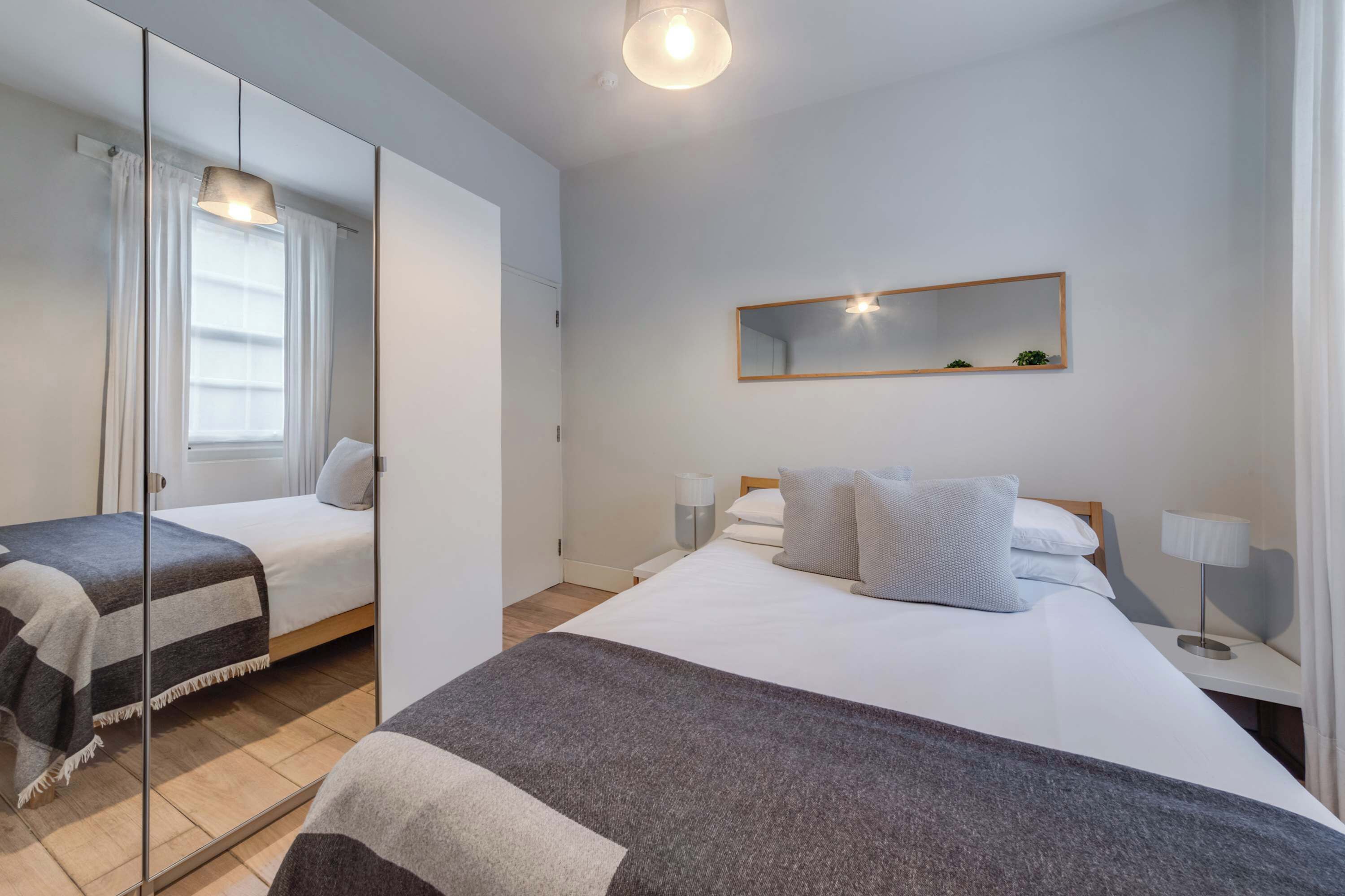 A modern hotel room with a large bed, a bedside table with a lamp, a mirror on the wall, and a window with curtains.