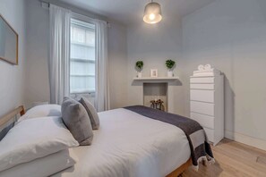 Un dormitorio con cama, mesita de noche, cómoda, ventana con cortinas y estante con objetos decorativos.