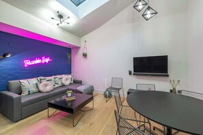 A modern living room with a sofa, coffee table, and dining area, featuring a neon sign and a flat-screen TV. - The Acre Abode - Covent Garden - by Frankie (London)