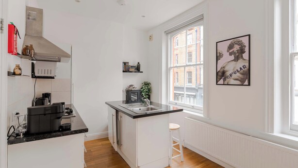 A compact kitchen with white cabinetry, a black countertop, a stainless steel range hood, a window with a view of buildings, and a framed picture on the wall. - Dionysus' Den - Fitzrovia - by Frankie Says (London)