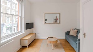 A modern living room with a large window, a round wooden coffee table, a blue sofa, and a mounted TV. - Dionysus' Den - Fitzrovia - by Frankie Says (London)