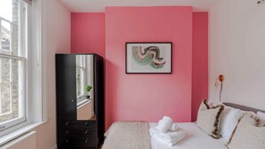 A bedroom with a pink wall, a bed with white bedding, a black wardrobe, and a framed abstract painting. - Dionysus' Den - Fitzrovia - by Frankie Says (London)