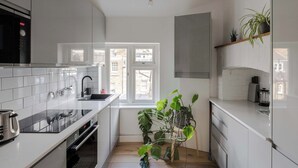 A modern kitchen with white cabinets, a black microwave, a sink, and a window with a view of buildings outside. - Stairway To Heaven - Fitzrovia - by Frankie Says (London)