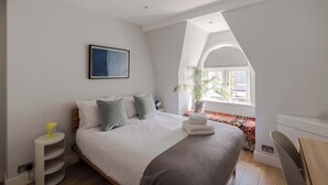 A bedroom with a bed, a bedside table, a potted plant, and a window with a view. - Stairway To Heaven - Fitzrovia - by Frankie Says (London)