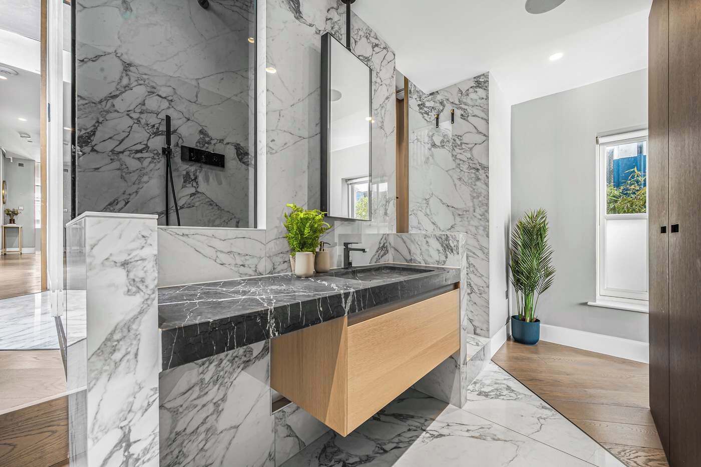 A modern bathroom with marble walls, a wooden vanity, a large mirror, and a potted plant.