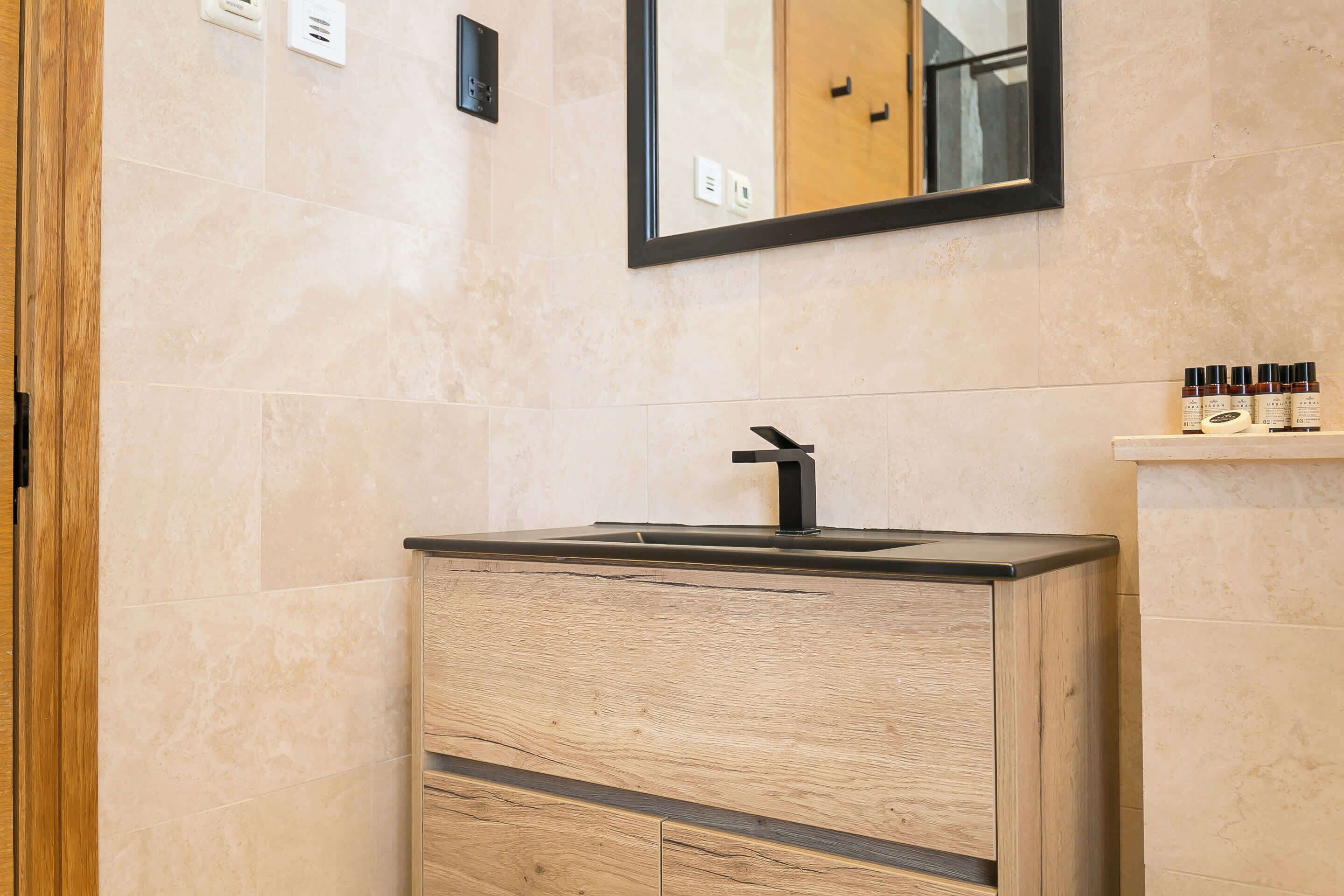 Une salle de bain avec un meuble-lavabo en bois, un robinet noir et un miroir au-dessus. On peut voir des articles de toilette sur une étagère à droite.