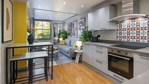 A modern kitchen with a built-in oven, a dining area with a table and chairs, and a sofa. - The Bloomsbury - by Frankie (London)